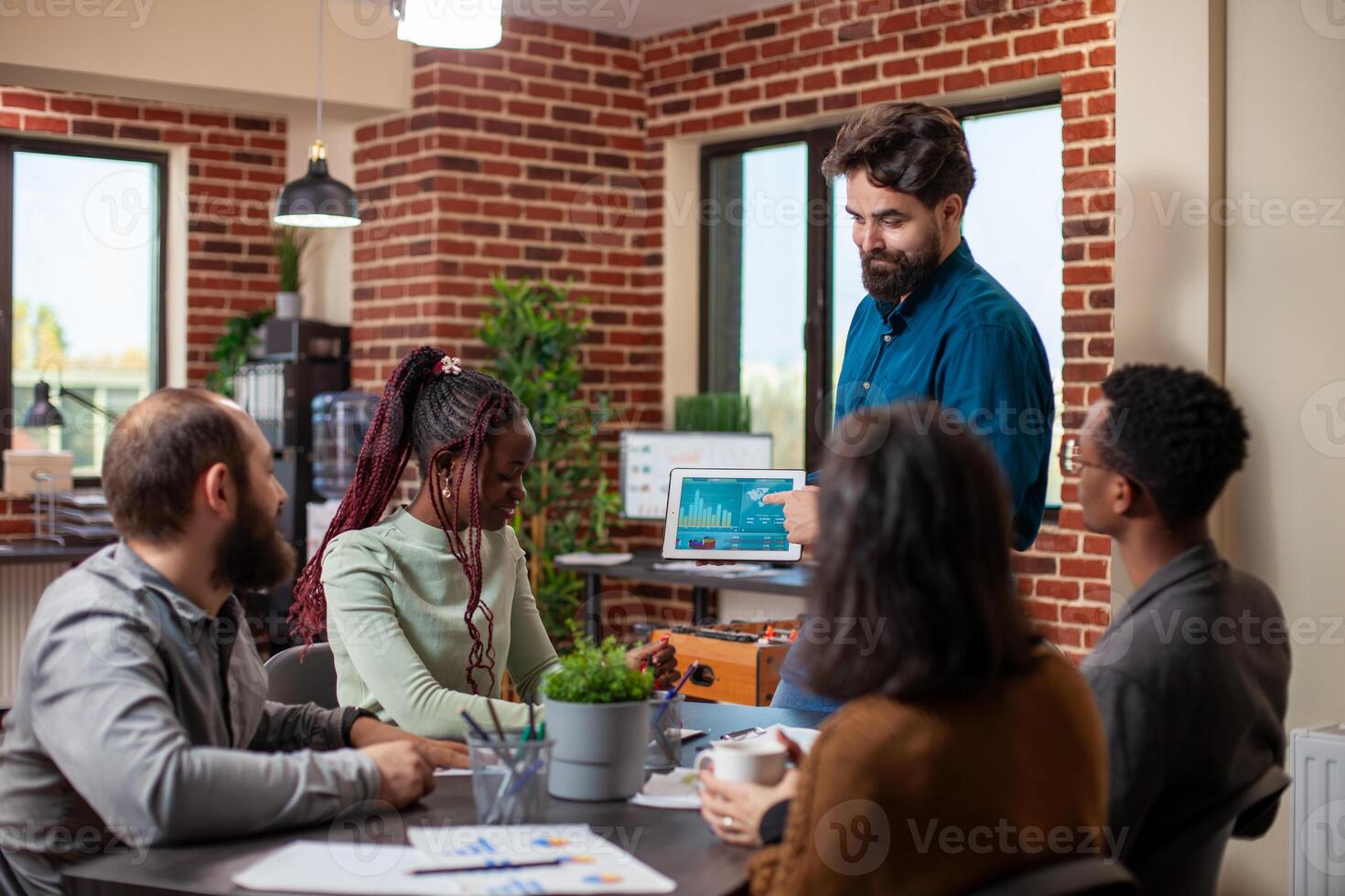 Caucasian manager holding tablet and showing financial graphs to his team seated at table in brick wall office. Bearded businessman using digital device, explaining company growth to his colleagues. photo