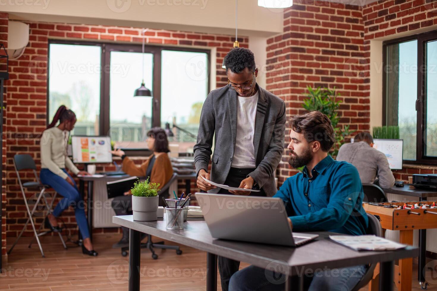 Male professional holds documents and shares ideas with his manager, brainstorming strategies in brick wall workspace. African american analyst with papers in hand, gives feedback to bearded coworker. photo