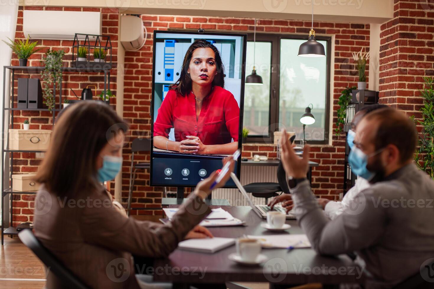Colleagues in brick wall office hold safe, masked virtual meeting with female client on tv screen, sharing strategy ideas. Woman manager on call on monitor speaking with team seated in boardroom photo