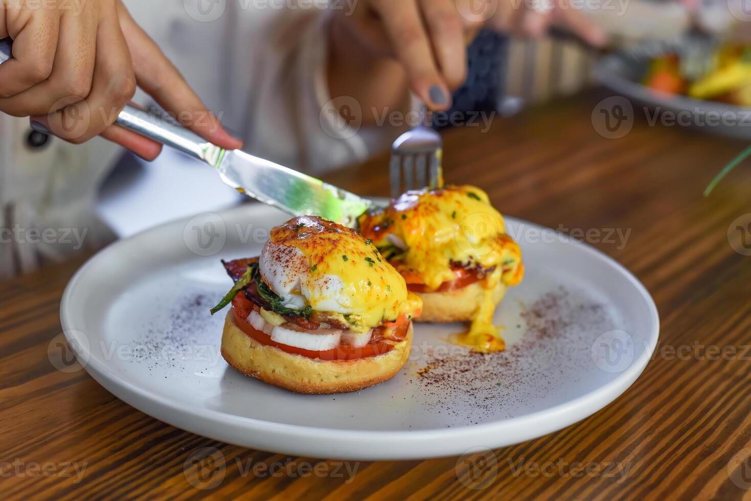 Hands with knife and fork cutting english muffins with poached eggs, bacon and Hollandaise sauce with vegetables photo