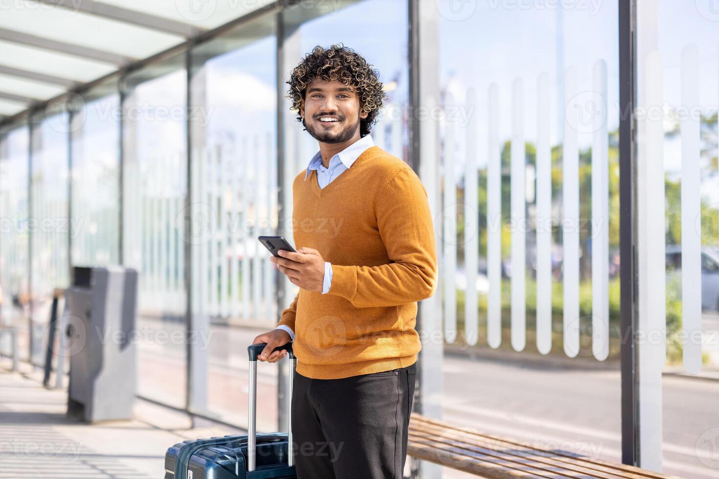 Portrait of a male passenger at a public transport stop, looking at the camera and smiling with a phone in his hands and a white travel suitcase. photo