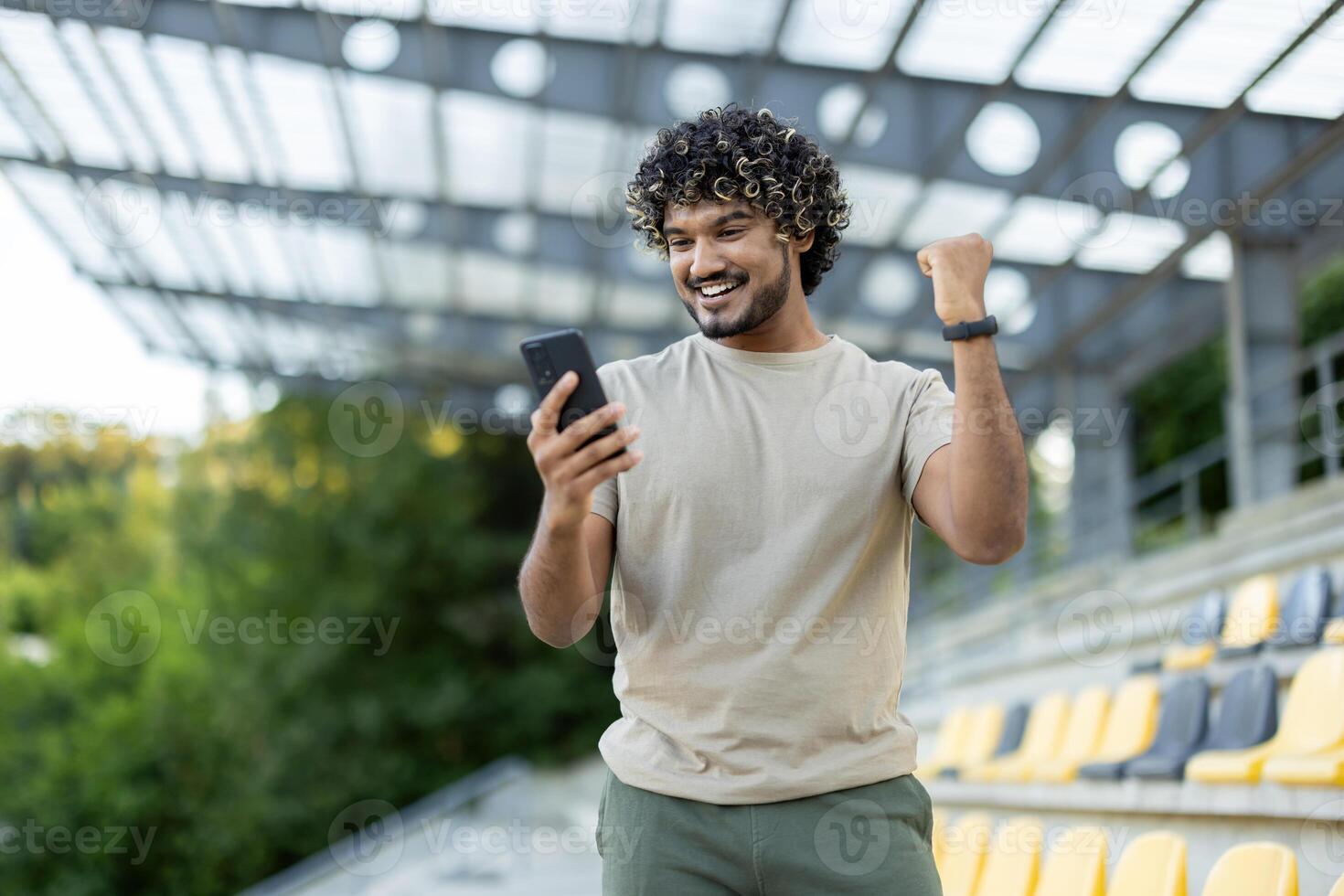 Man in stadium holding phone in hands, satisfied with training results, holding hand up celebrating. Athlete checking app for online training and recording achievement results. photo
