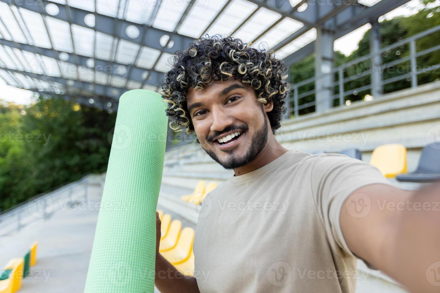 A man takes a selfie and talks to his friends using a call app on his phone. The athlete holds a fitness and yoga mat in his hands.. photo