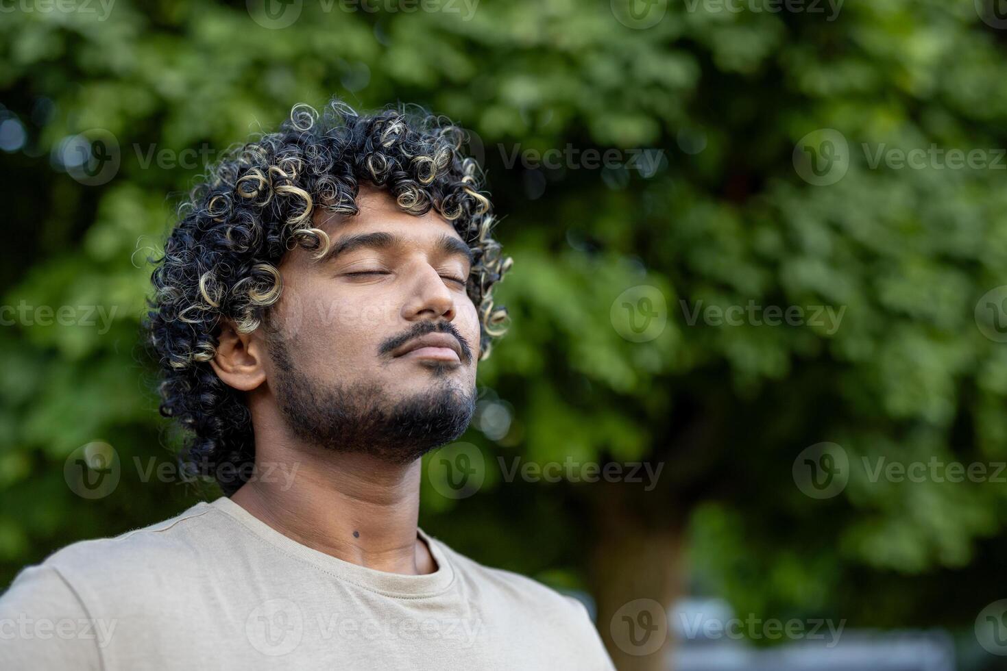 A serene portrait of a man with curly hair enjoying a peaceful moment outside surrounded by green leaves, highlighting relaxation and mindfulness in nature photo