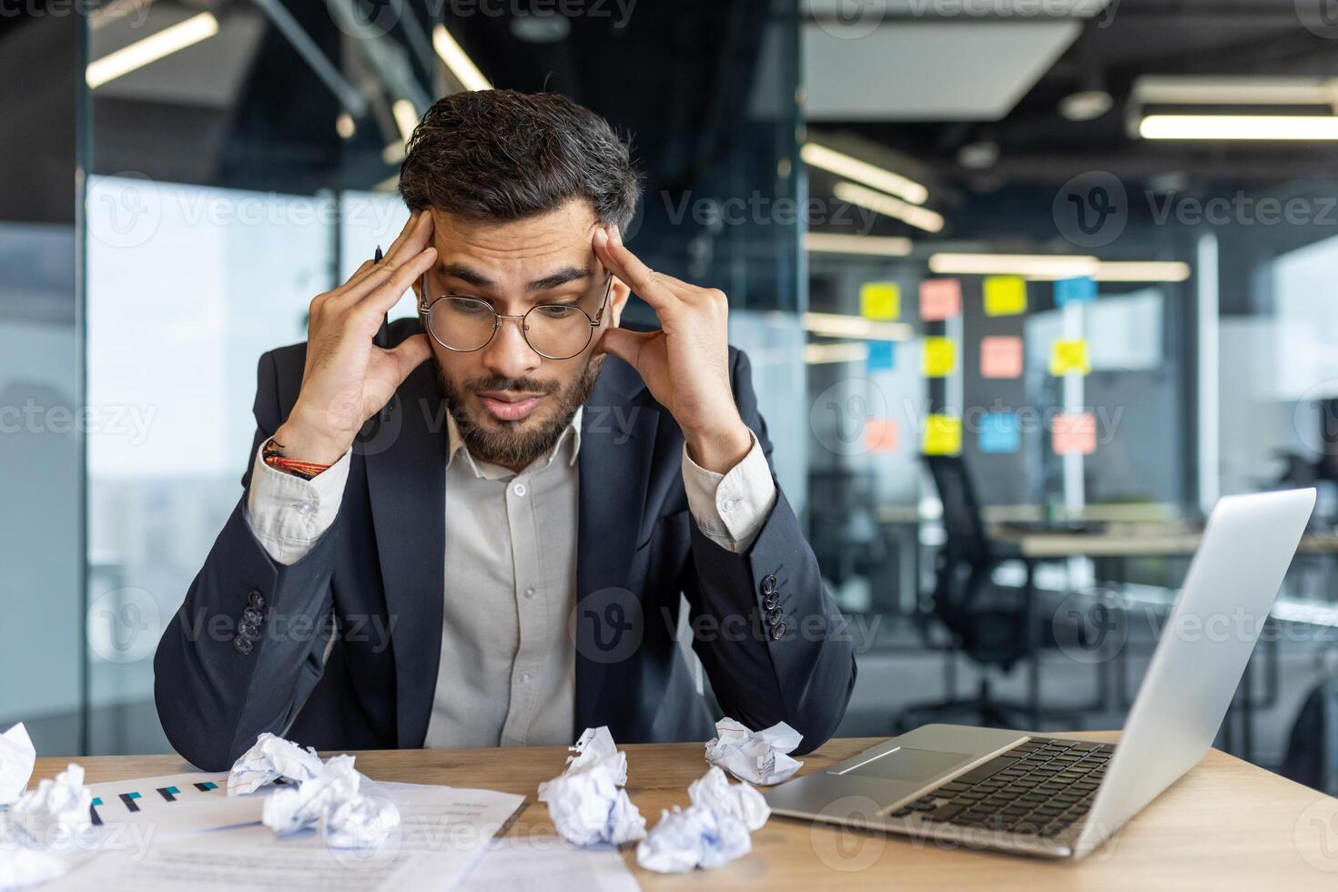 Frustrated and overwhelmed man sitting at workplace inside office, unsuccessful financial report. Businessman dissatisfied with financial results, near scraps of paper. photo