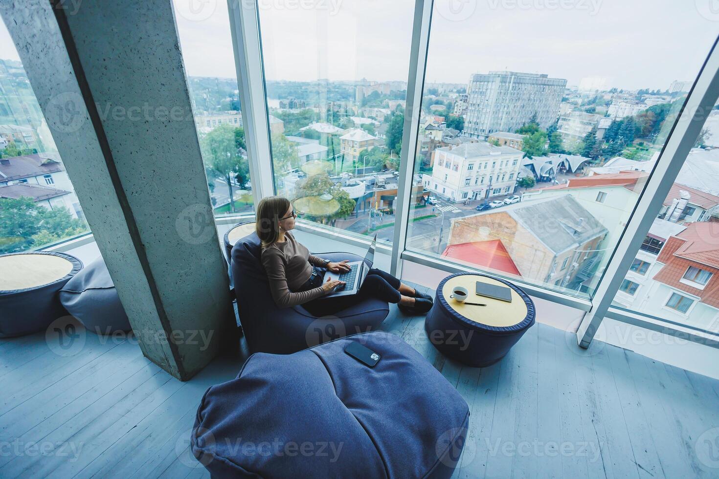 A beautiful female student works on a laptop while sitting on a bean bag chair in a modern creative office against the background of panoramic windows. Break time concept photo