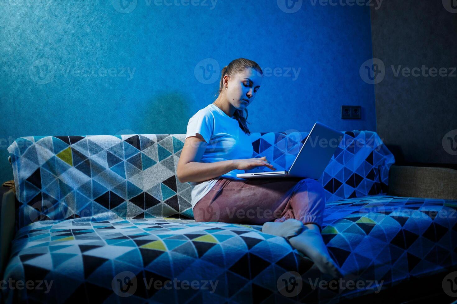 A girl in a white T-shirt is sitting at home on the couch with a laptop on her lap photo