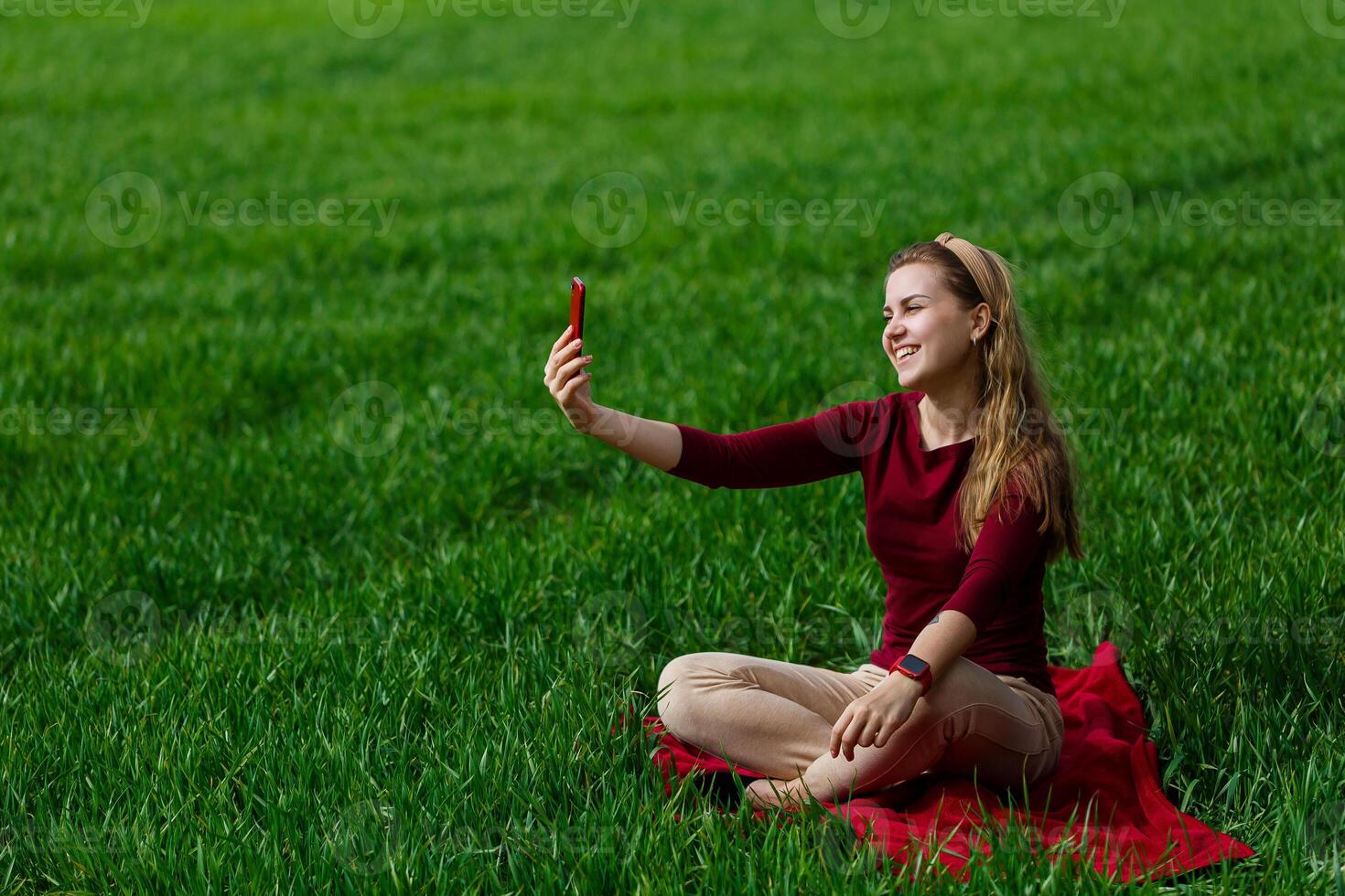 Beautiful woman with a phone in her hands sits on the grass. The girl looks at herself in the camera of the phone and takes selfie photos. She smiles and enjoys a warm day. Concept photo on smartphone
