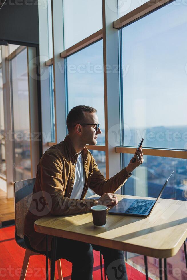 happy young man wearing glasses and casual clothes is talking on the phone while working on a laptop from a cozy workspace. A successful freelancer works remotely photo