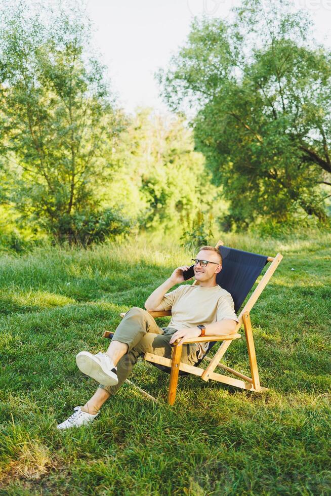 A man is talking on a smartphone while sitting on a bench in a city park. A young guy reads the news, writes a message on his mobile phone. Recreation in nature, remote work on vacation photo