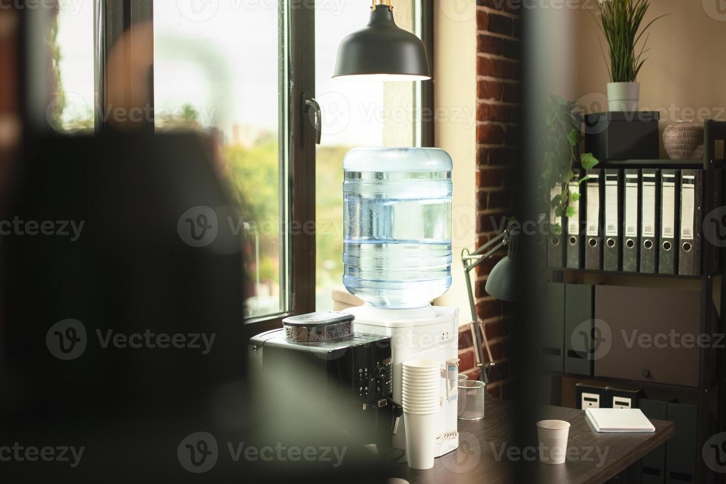 Corner office space having coffee machine, water cooler, and plastic cups positioned beside folder cabinet. Refreshments and file binders neatly arranged in brightly lit brick wall room. photo