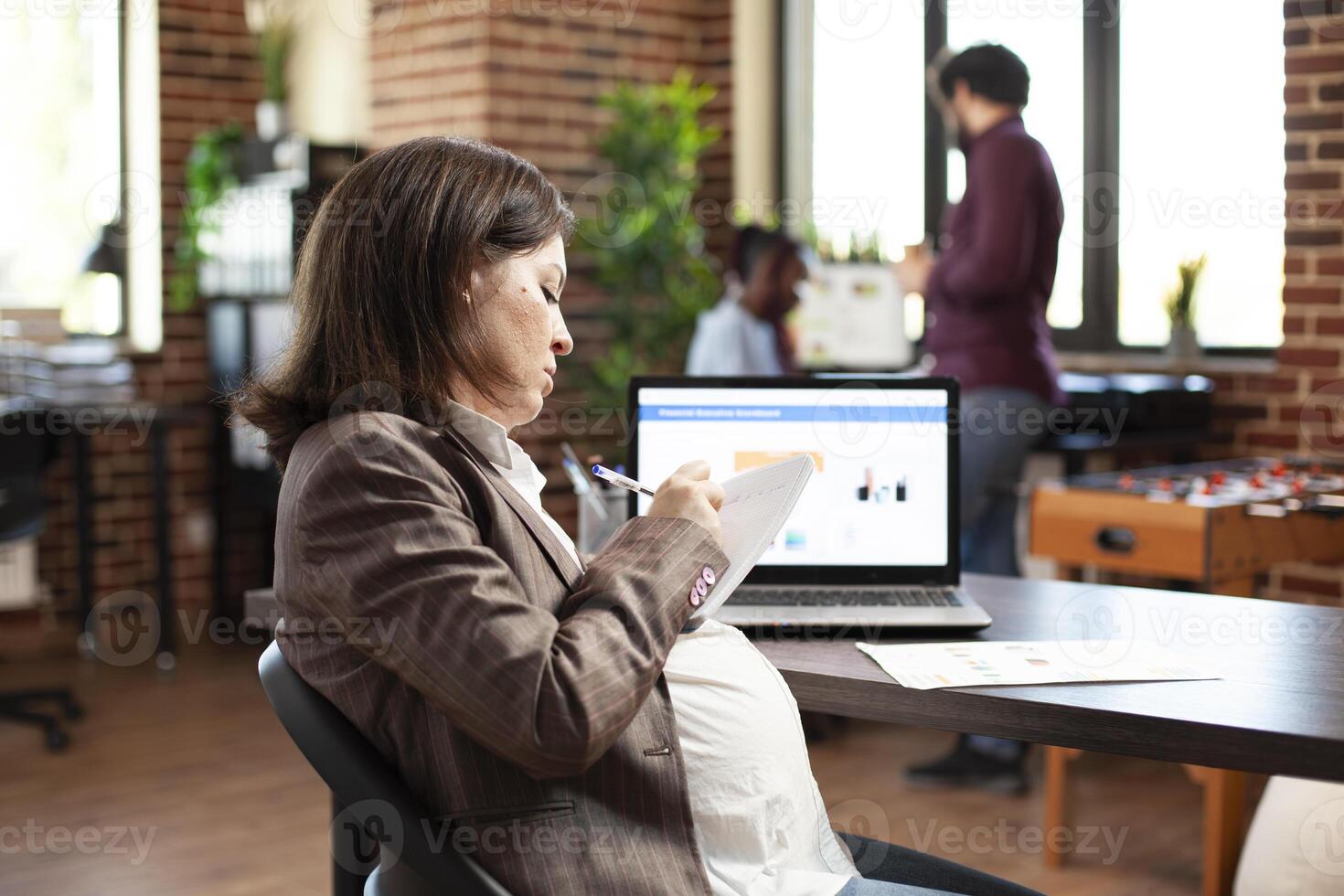 Young expectant mother seated at desk, writing research data on her notepad. Pregnant white woman with book is working in startup company room, having laptop opened on table. photo
