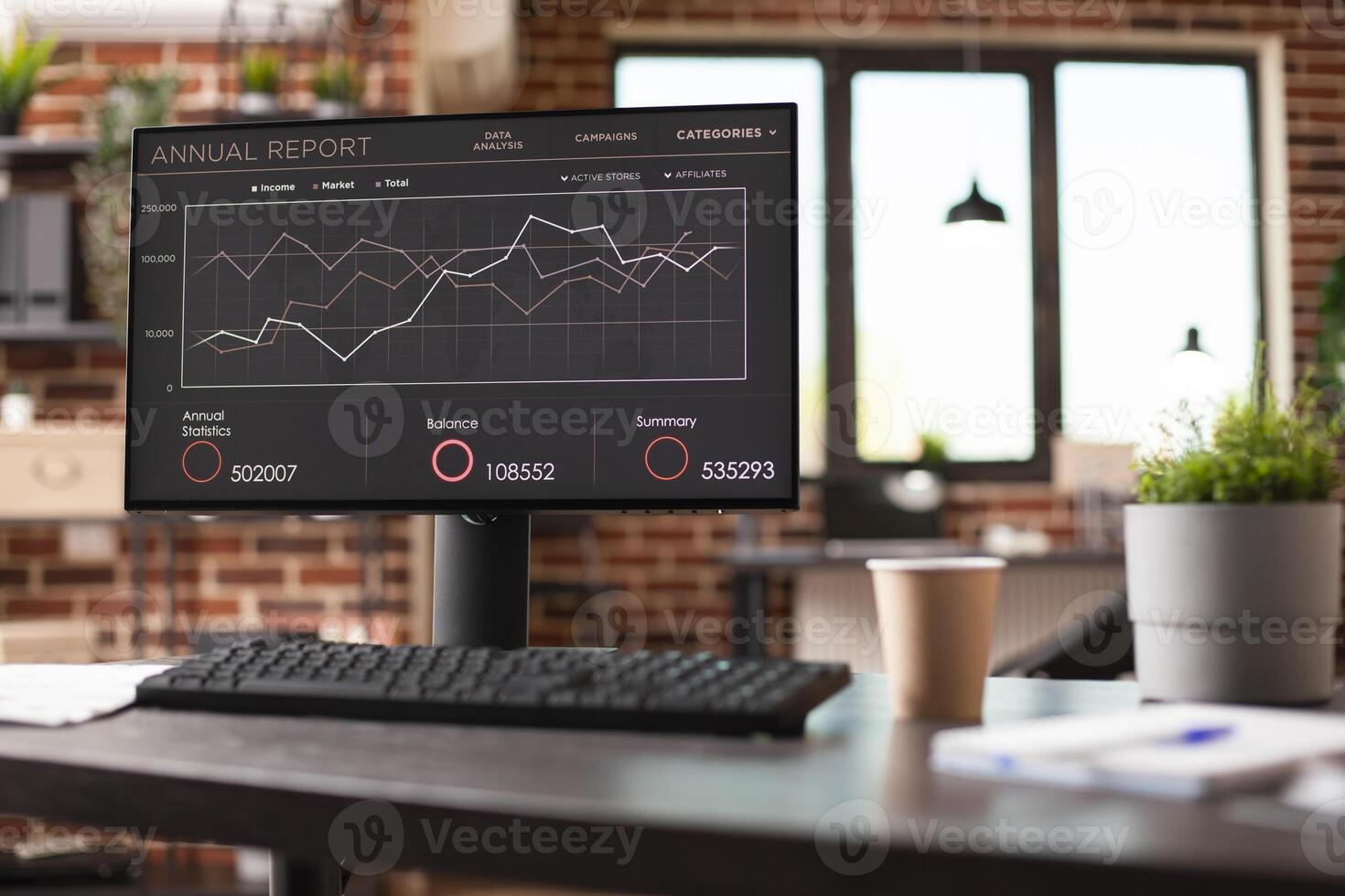 Computer screen displaying company analytics and financial data sits on modern desk in vacant brick wall office. Indoor plant and file folders placed on table, add professional touch. photo