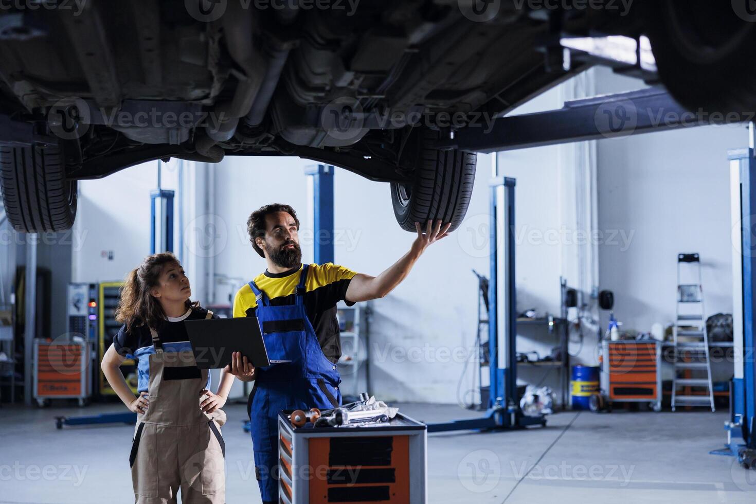 Engineer in car service uses laptop to calculate invoice after fixing broken automobile wheels. Garage worker uses device to inform client of final costs after mending vehicle on overhead lift photo