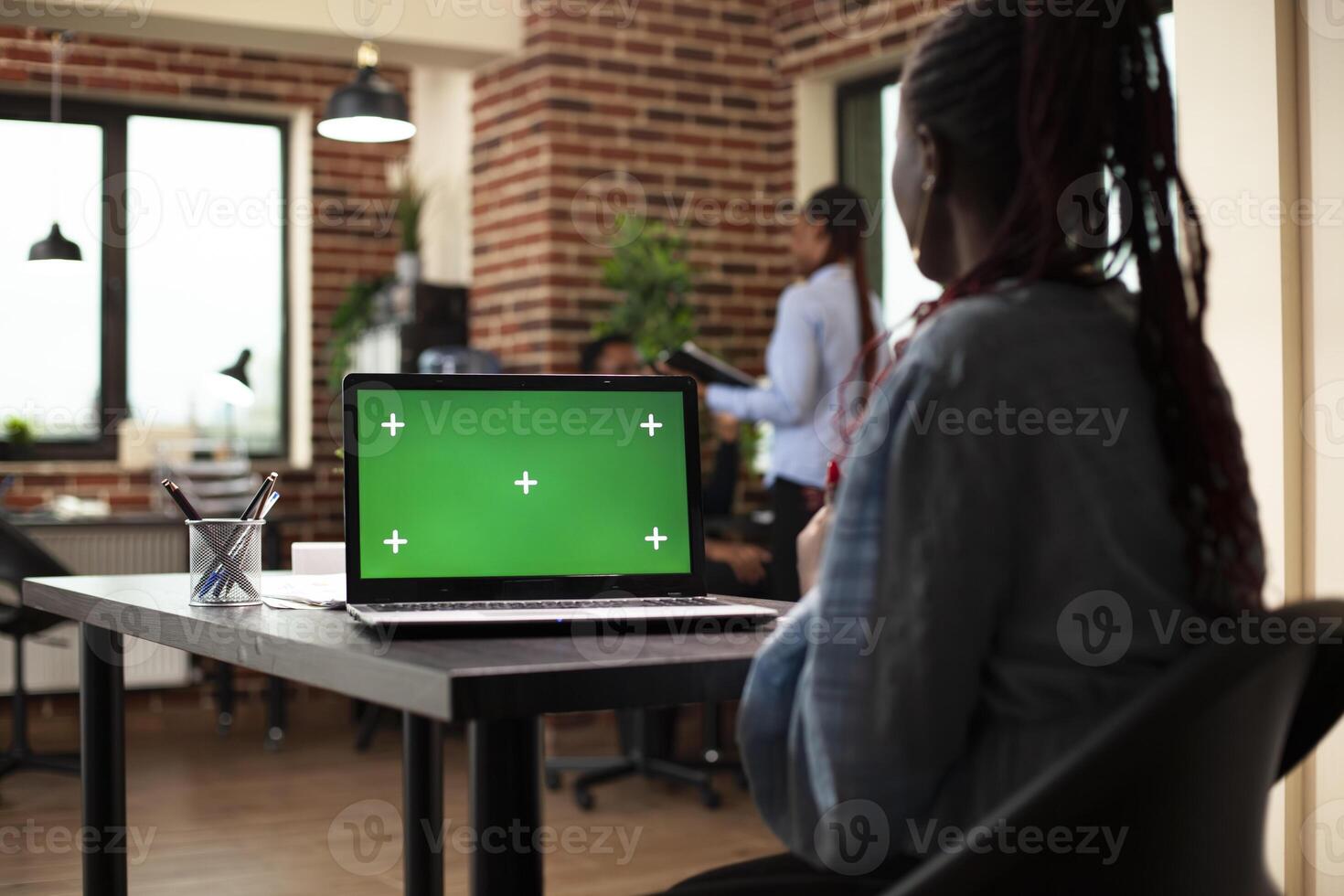 Black businesswoman seated at desk, using laptop with a greenscreen display. African american female manager looks at device screen displaying isolated chromakey mockup template in office. photo