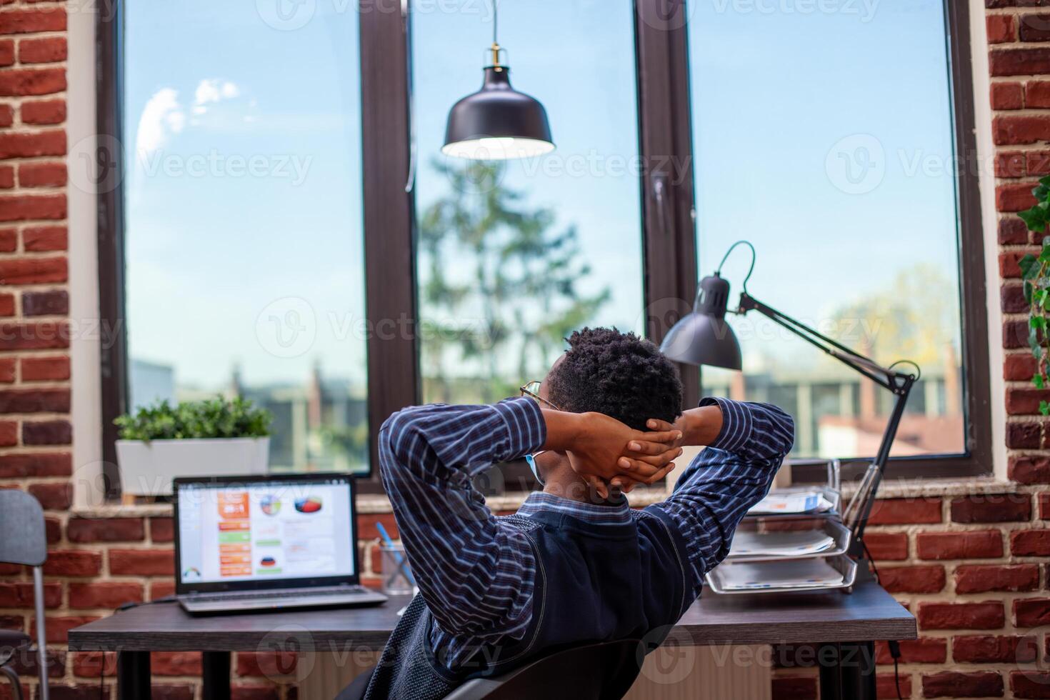 Businessman leans in office chair, hands resting behind his head, stretching and taking a break after busy schedule. African american male manager relaxing at his desk, feeling satisfied after work. photo