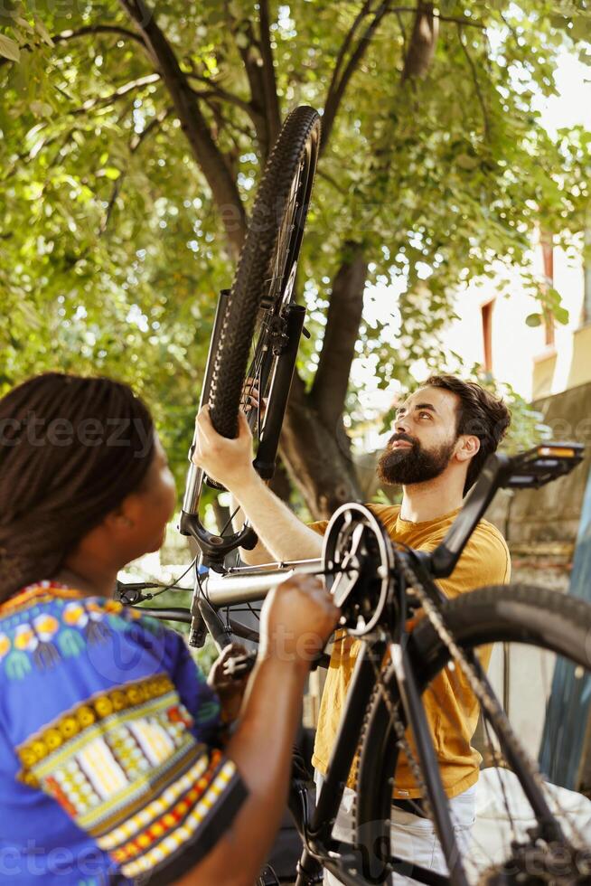 Young multiracial pair fixes damaged bicycle outside using professional equipments. Healthy active african american woman and causian man inspecting and maintaining bike tires and chains for summer cycling. photo