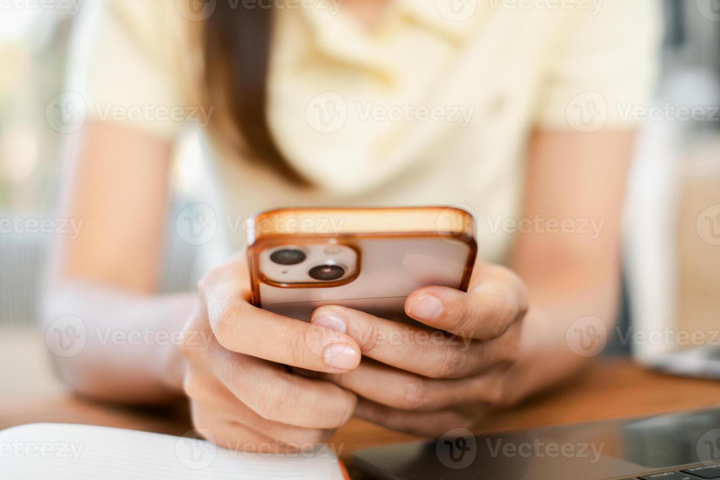A person holding a smartphone with an orange case, seated at a wooden table, focusing on the screen. photo