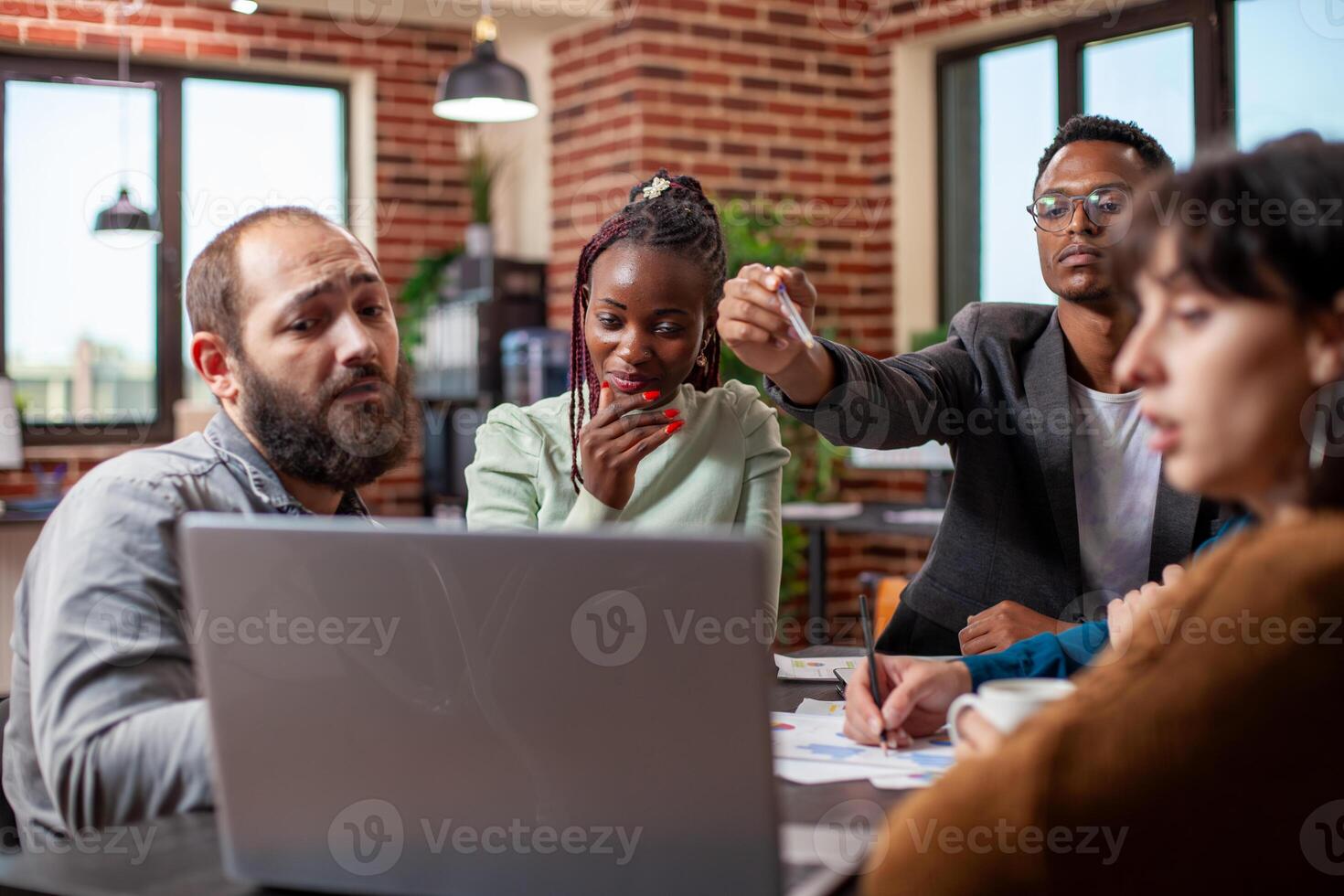 Small group of employees collaborating around a laptop, displaying teamwork in modern office. African american and caucasian colleagues look at device screen, focused on startup project discussions. photo