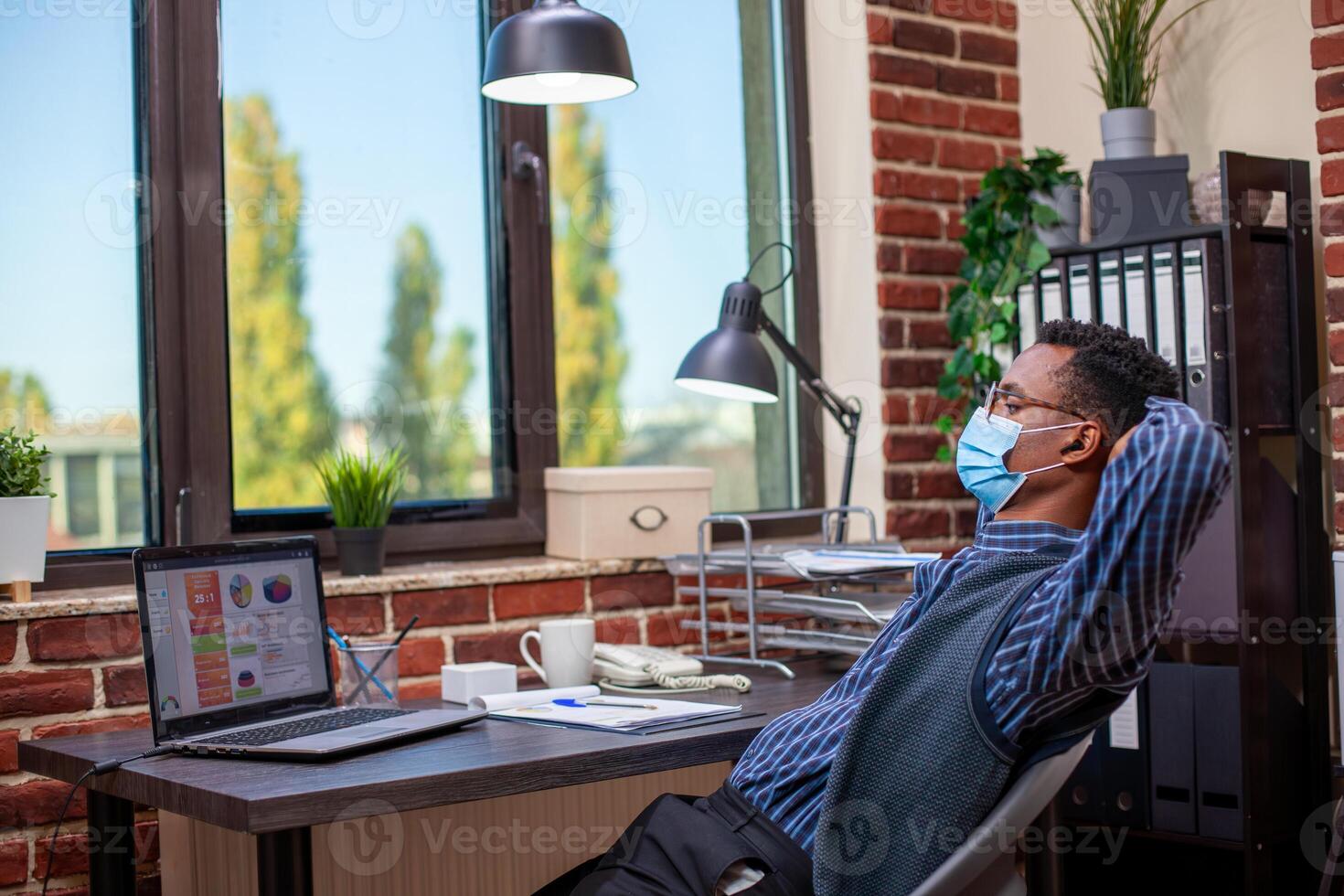 Professional analyst with face mask, leaning back in chair and unwinding in brick wall workspace. Businessman looking at laptop screen with hands behind his head, enjoying a break in startup office. photo