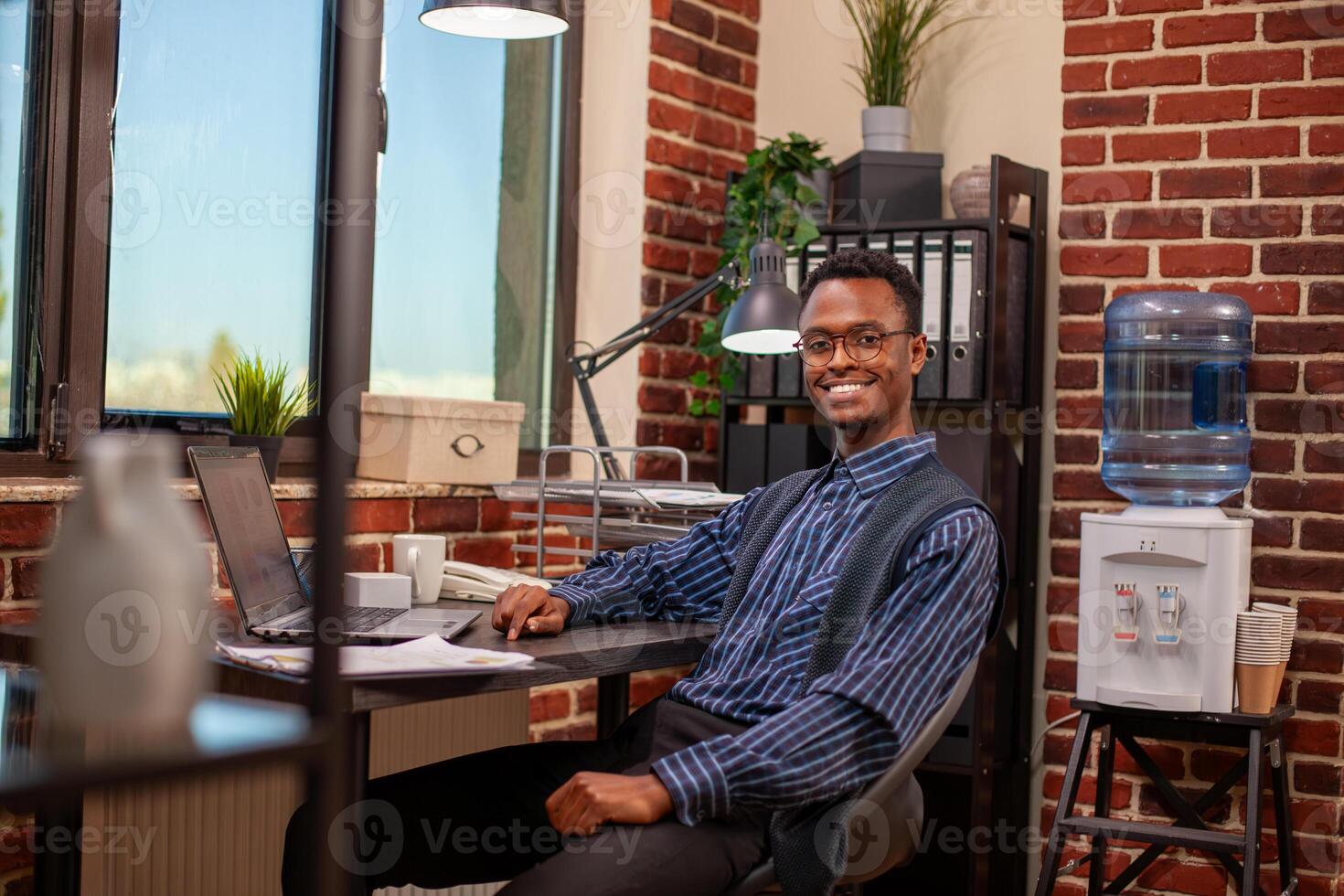 Portrait of black male analyst sitting at neatly arranged desk, looking directly at camera with confidence. Professional man poses with cheerful smile in office, having laptop and documents nearby. photo