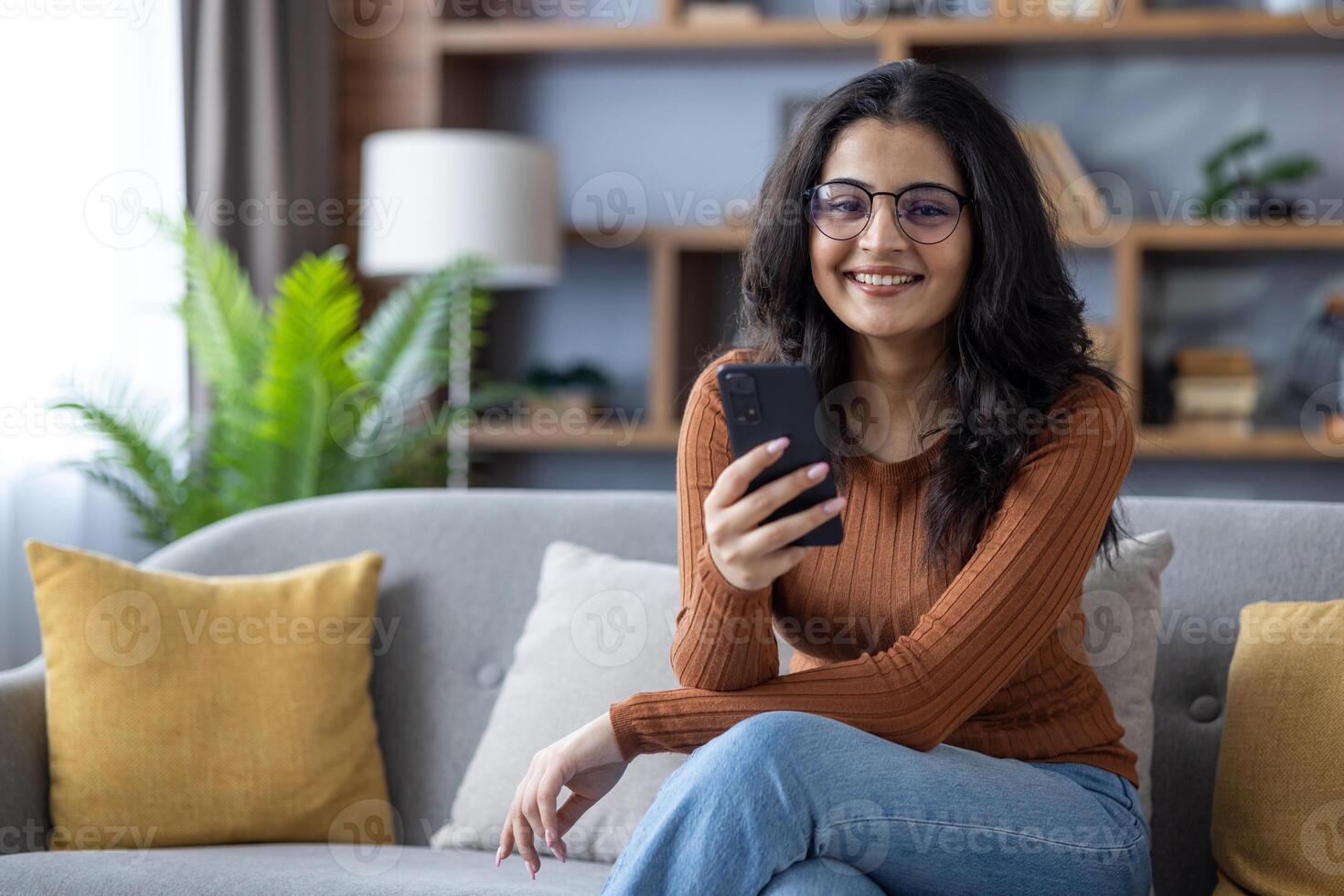 Portrait of a smiling young Indian woman in glasses sitting on the sofa at home, holding a mobile phone in her hand, looking at the camera. photo