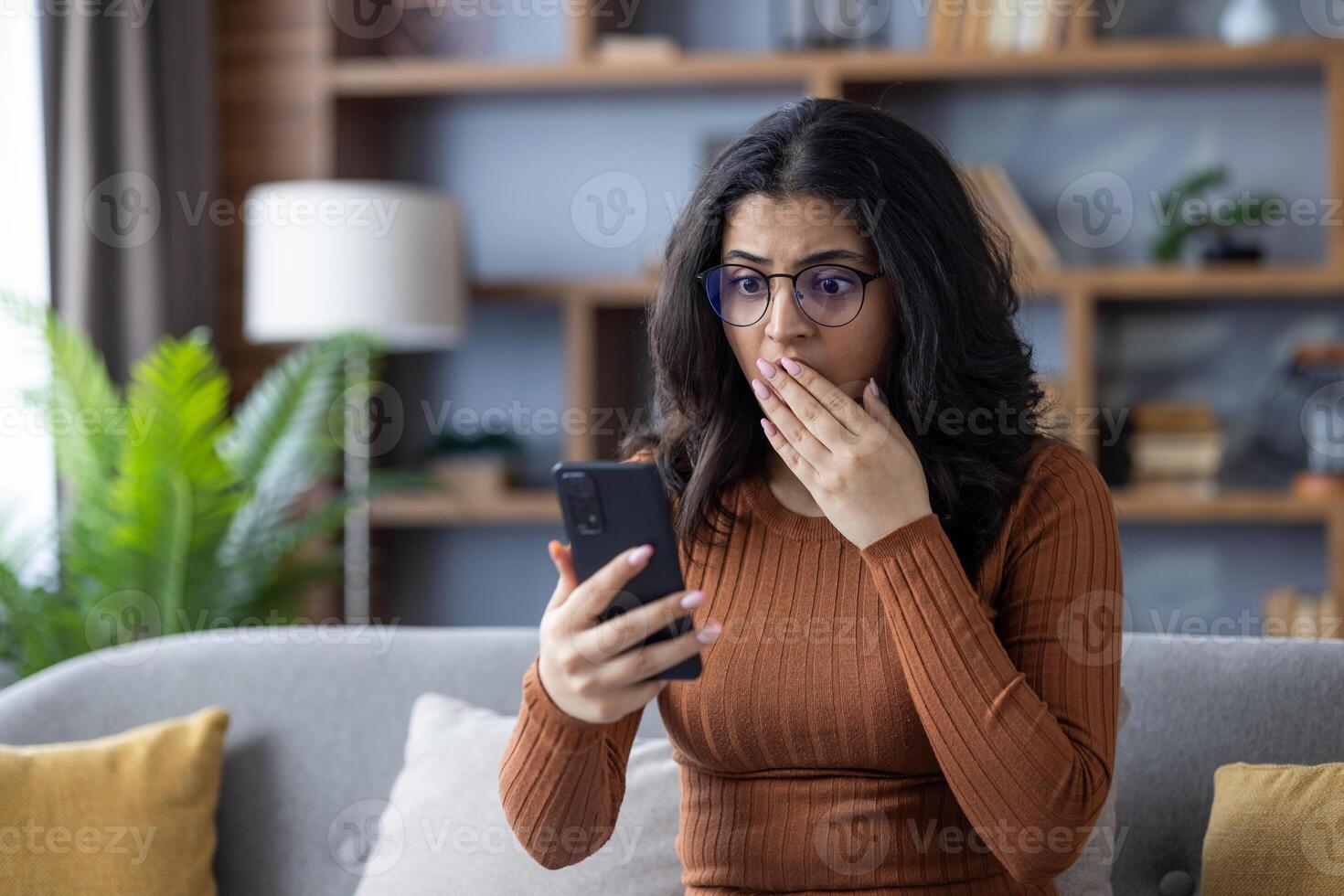 Close-up photo of a sad young Indian woman sitting on the sofa at home, looking at the phone in shock, covering her mouth with her hand.