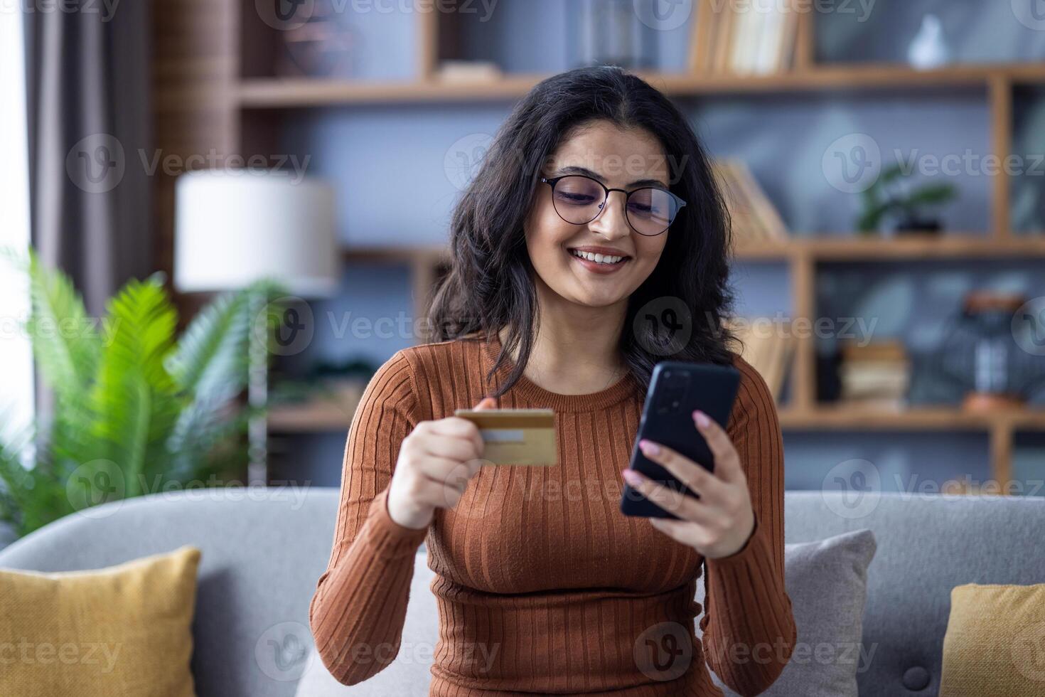 Close-up photo of a young smiling Muslim girl at home, sitting on the sofa, holding a credit card and using a mobile phone.