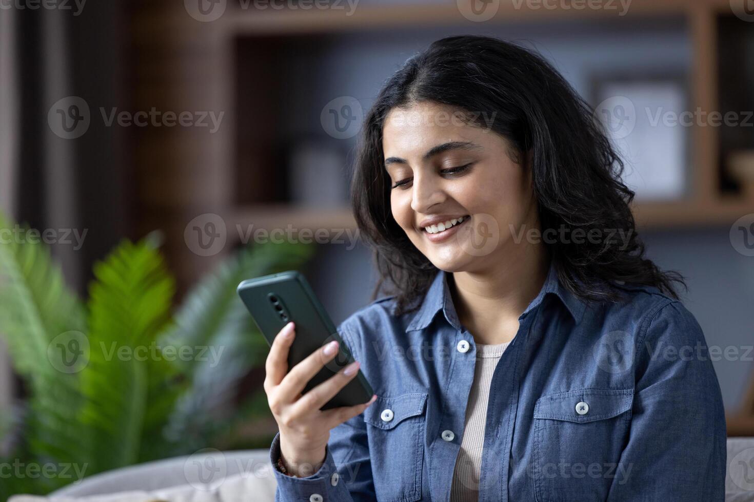 Close-up photo of a young Indian woman in a denim shirt at home, sitting on a sofa and using a mobile phone with a smile.