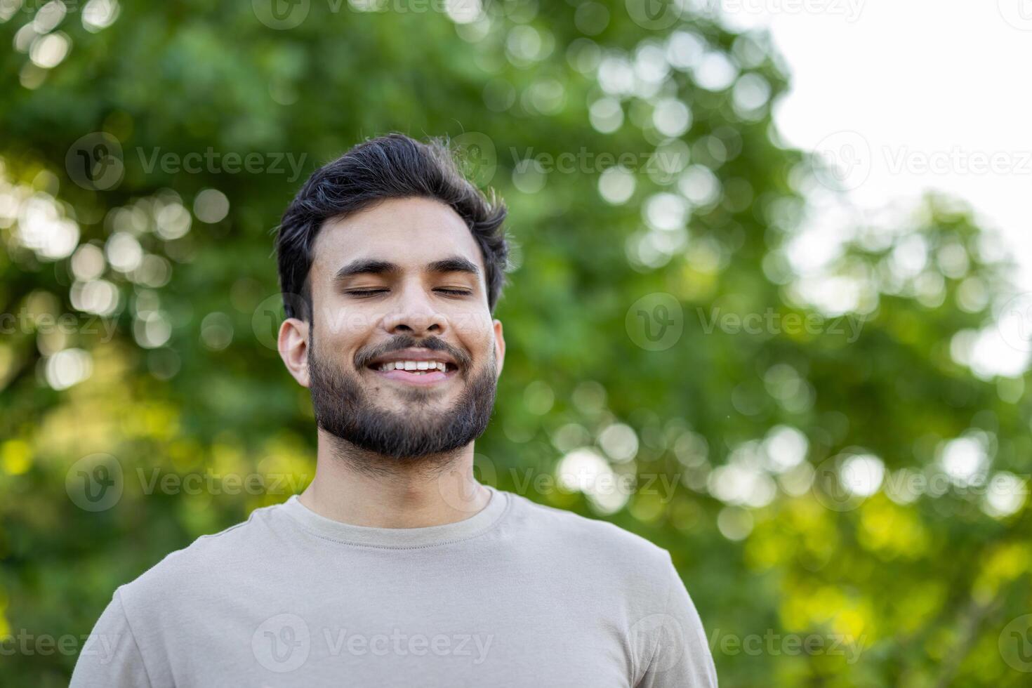 Close-up photo of a happy and satisfied young Indian man standing in a park with his eyes closed, resting and breathing deeply.