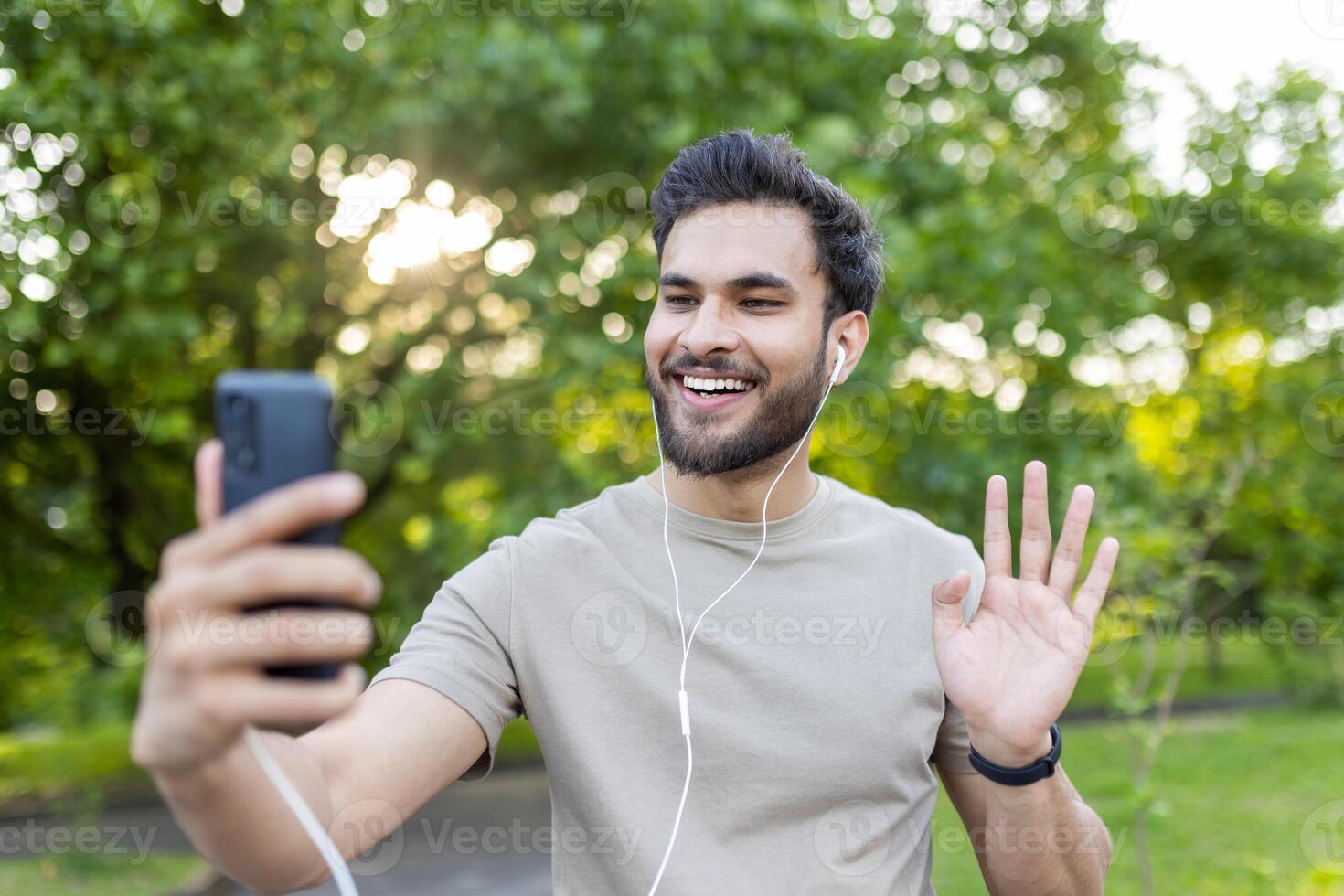 Close-up photo of a young Indian man in headphones doing sports and jogging in the park, talking on the phone on a call, waving at the camera.