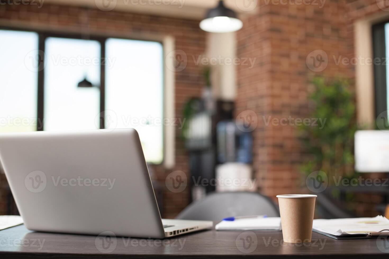 Empty brick wall workspace having table holding laptop, coffee cup and company documents, ready for busy workday. Startup office with no people is well prepared for business meetings and research. photo