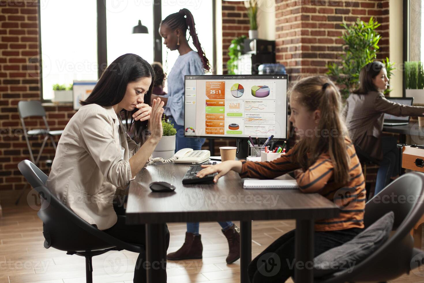 Working mom reviewing market strategy on desktop pc and annoyed by daughter interrupting at desk in startup office. Little girl plays with keyboard on table as woman is trying to work on computer. photo