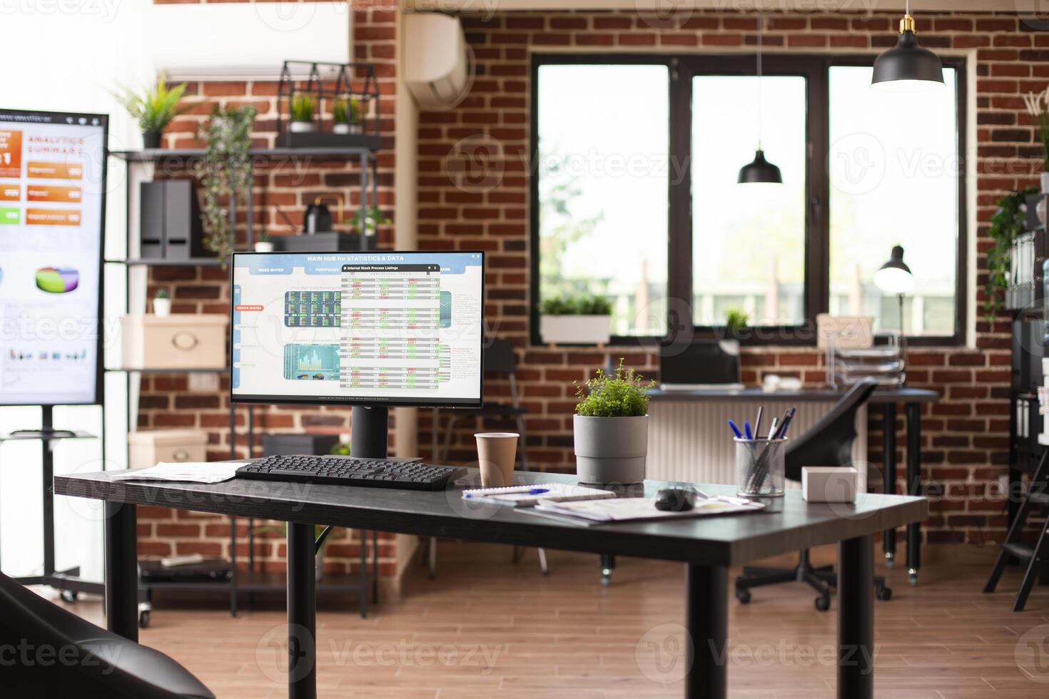 Indoor startup office displaying desktop pc with financial data on well organized desk. Brick wall workspace includes modern technology, shelves, and furniture arranged, ready for employees to use. photo