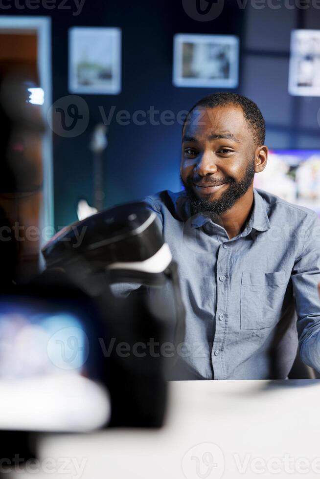 Smiling african american man presenting VR goggles to audience, filming with professional camera. Content creator trying virtual reality headset product before recommending it to fans photo