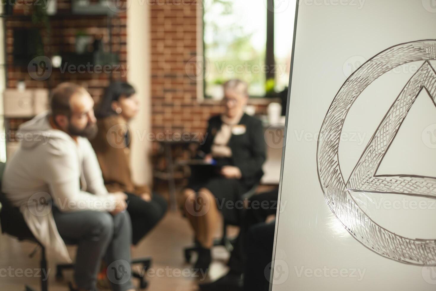 Closeup of aa meeting symbol on white board with ongoing therapy session in background. People engaging in discussions about alcohol addiction with guidance from retired therapist in brick wall office photo