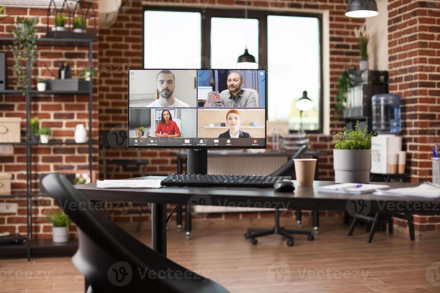 Vacant startup office with computer screen on clean desk shows virtual group training in progress. Shelves, water dispenser, and well organized file folders completes the modern brick wall workspace. photo