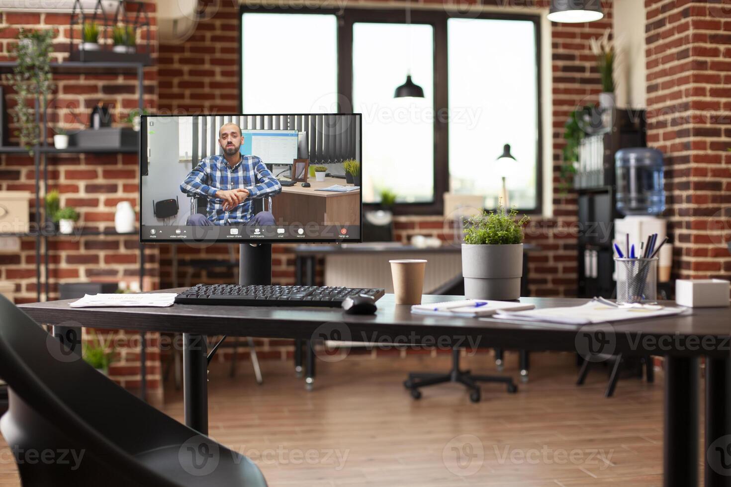 Desktop monitor on wooden table in well arranged room displays virtual meeting with male wheelchair user. Spacious room organized with computers and office supplies for effective planning. photo