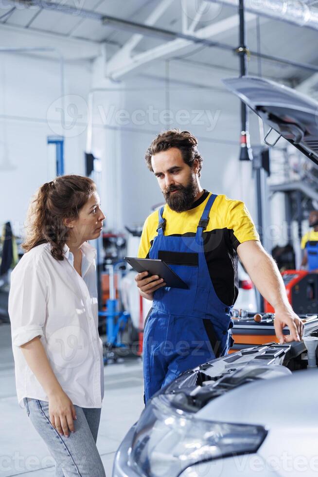 Mechanic in car service uses laptop to calculate invoice for client after repairing alternator. Friendly employee in garage with customer calculating final costs after servicing vehicle photo