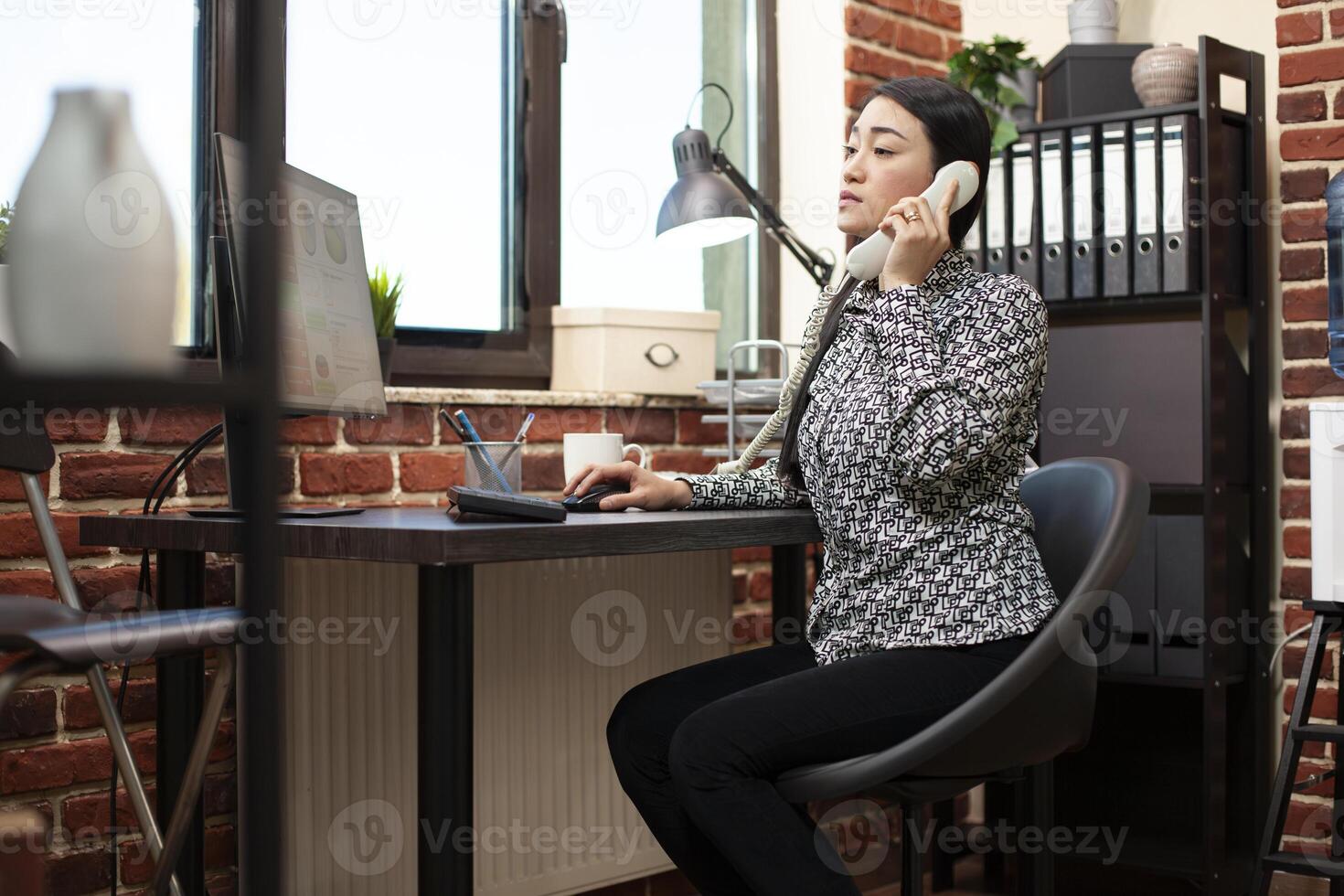 Financial advisor reviews analytics on computer, discussing project updates with a client over a phone call. Businesswoman references company documents during a landline conversation. photo