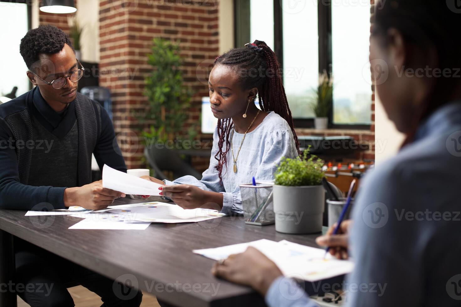 Male and female coworkers holding and reading documents at desk in modern workspace. African american colleagues sitting and reviewing financial paperwork during a meeting in startup office. photo