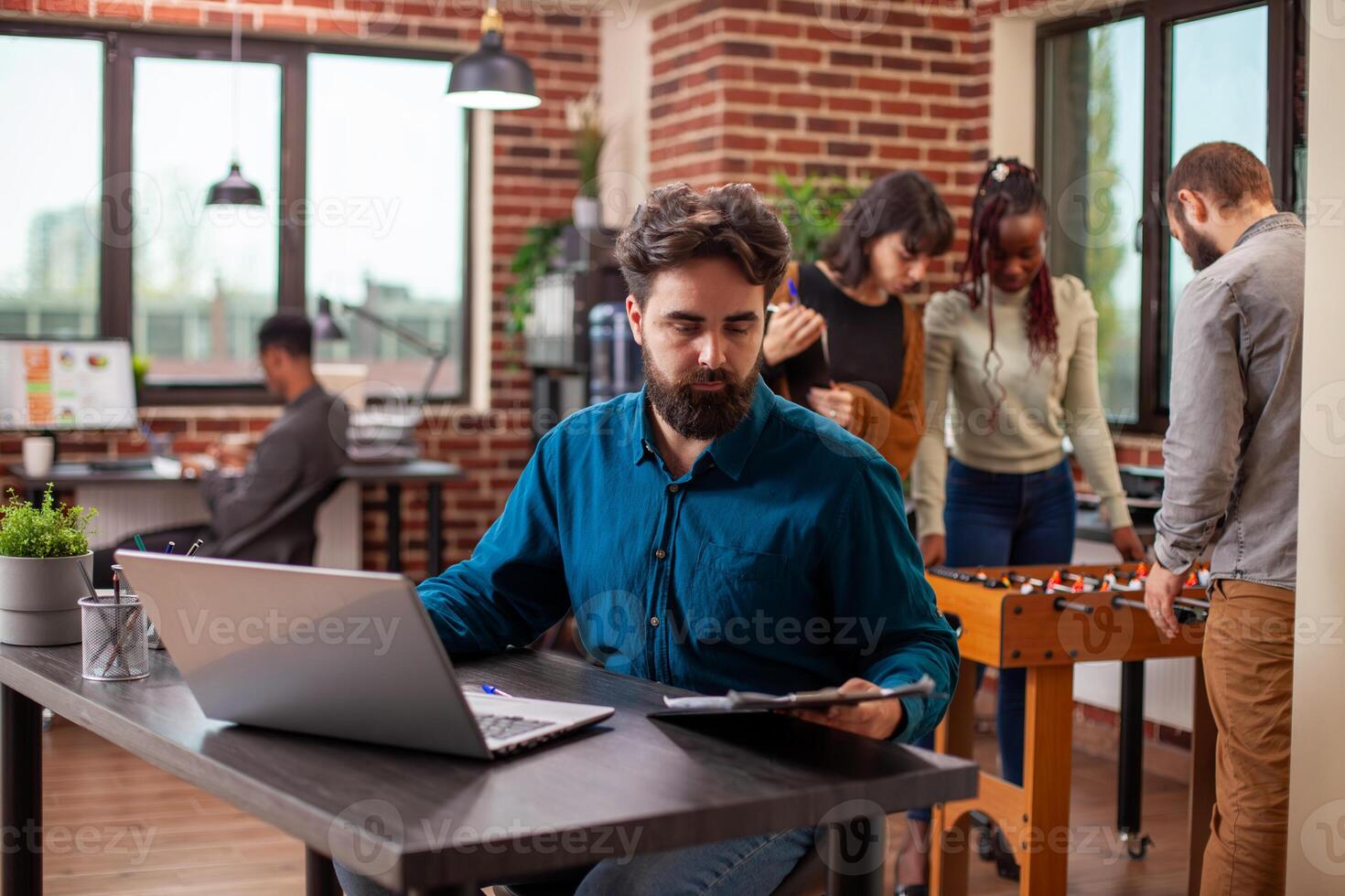 Male office worker focused on reviewing documents and charts on clipboard at desk, laptop placed beside him in brick wall workspace. Young man sitting at table and reading company guidelines. photo