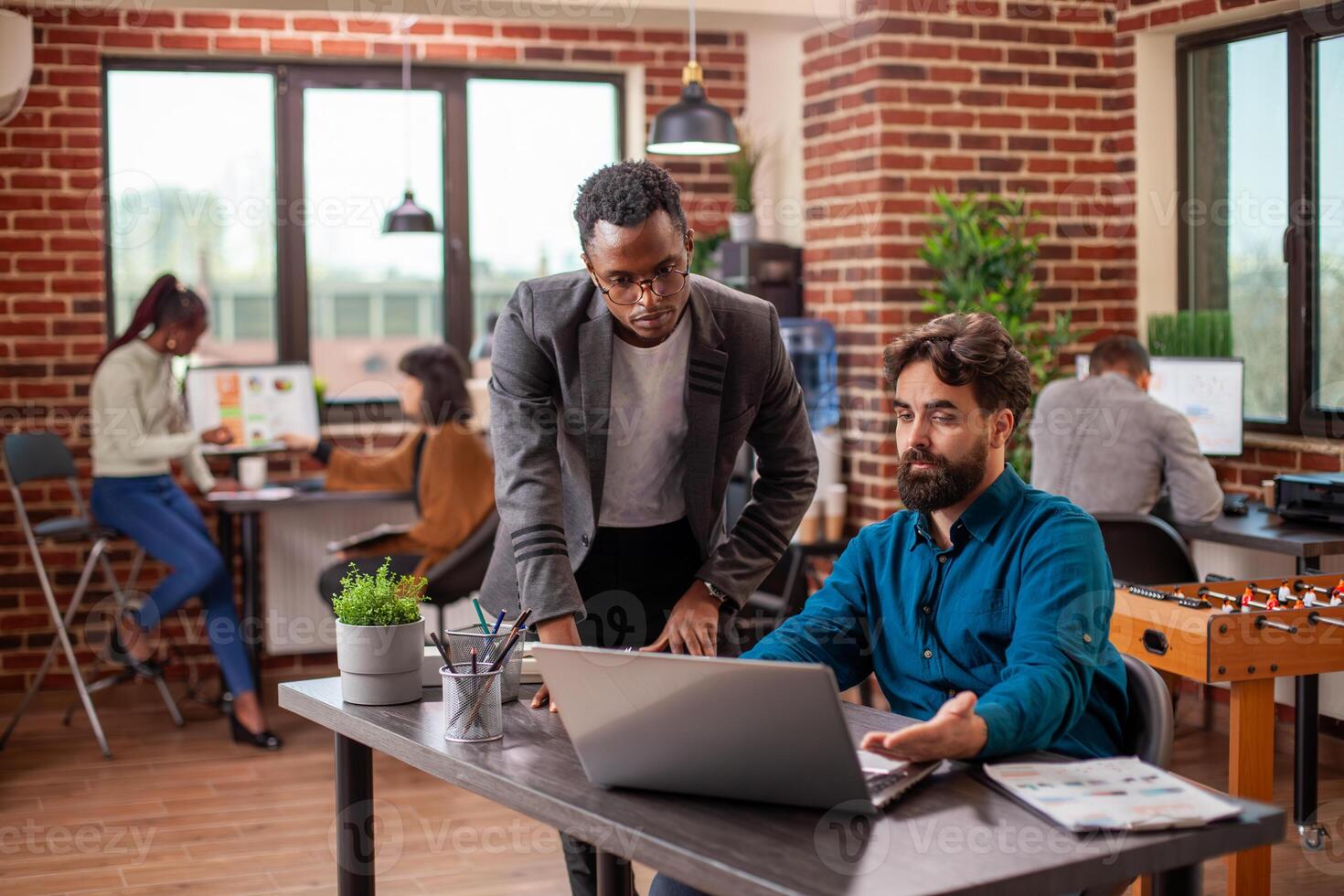 Caucasian man with laptop reviews online documents with african american coworker at table in modern workspace. Male colleagues remain focused on achieving business objectives using digital device. photo