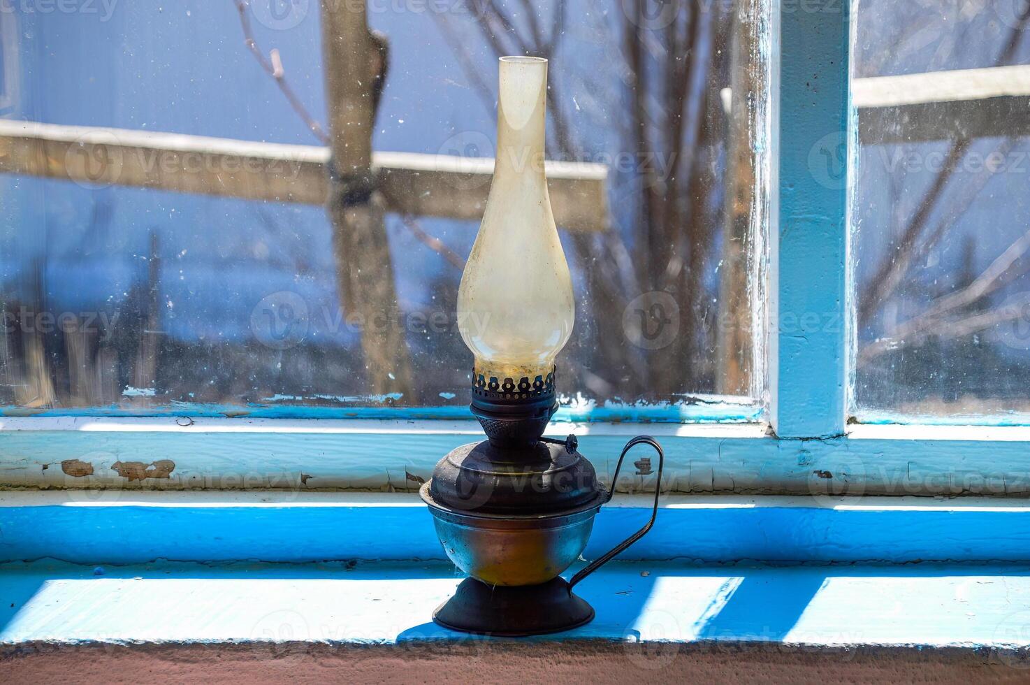 Used flat-wick kerosene lamp also known as a paraffin lamp on old shabby blue windowsill in the sunlight photo