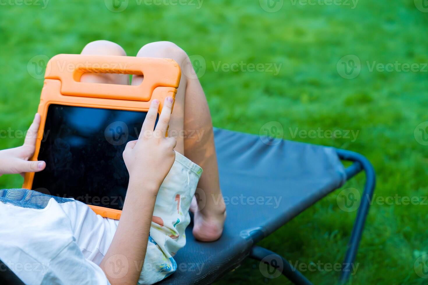 Child relaxing outdoors on a lounge chair using a tablet in an orange protective case photo