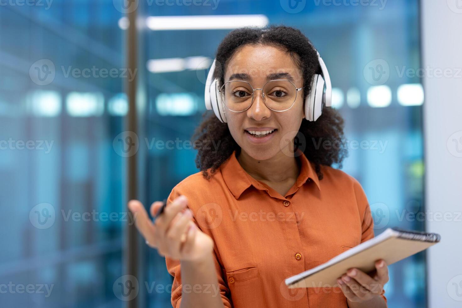 Webcam view, smiling woman in headphones and with a notebook in hands looking at camera, watching an educational course. Businesswoman inside the office on a remote business meeting conference. photo
