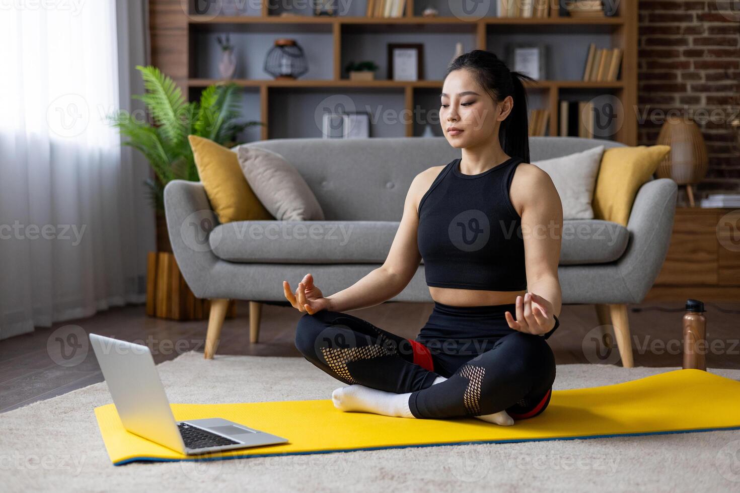 Asian woman practices yoga meditation at home on yellow mat, following routine on laptop, promoting relaxation, well-being, mindfulness. Peaceful indoor workout setting enhances focus, tranquility. photo