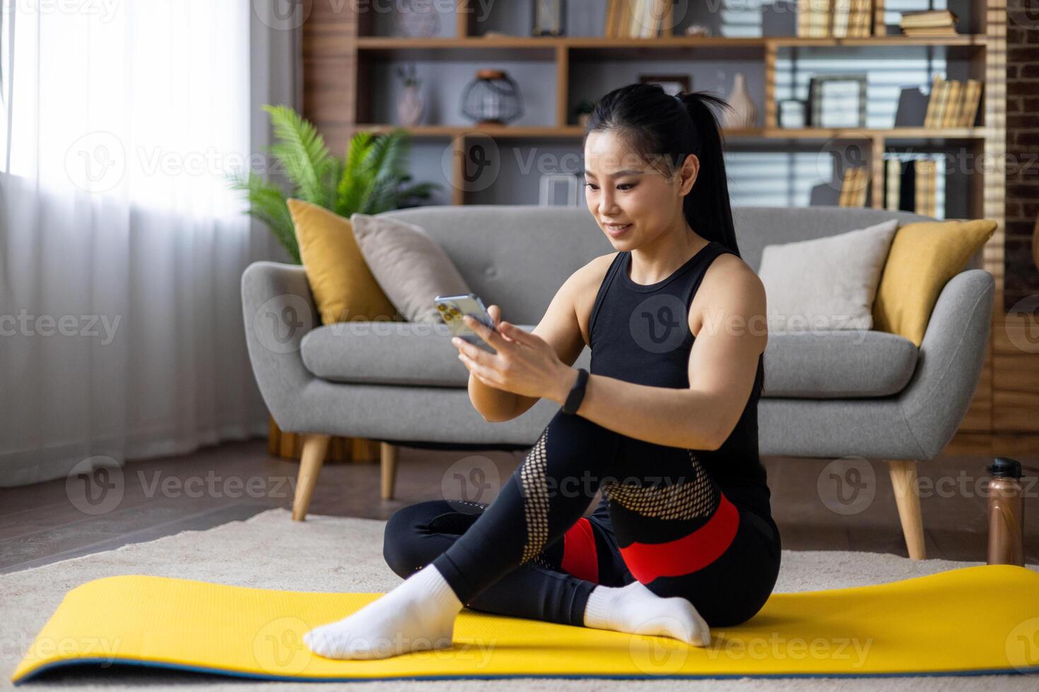 Asian woman sitting on yoga mat, using smartphone for fitness app or guided exercise. Dressed in athletic wear, she smiles, reflecting motivation and relaxation in home environment. photo
