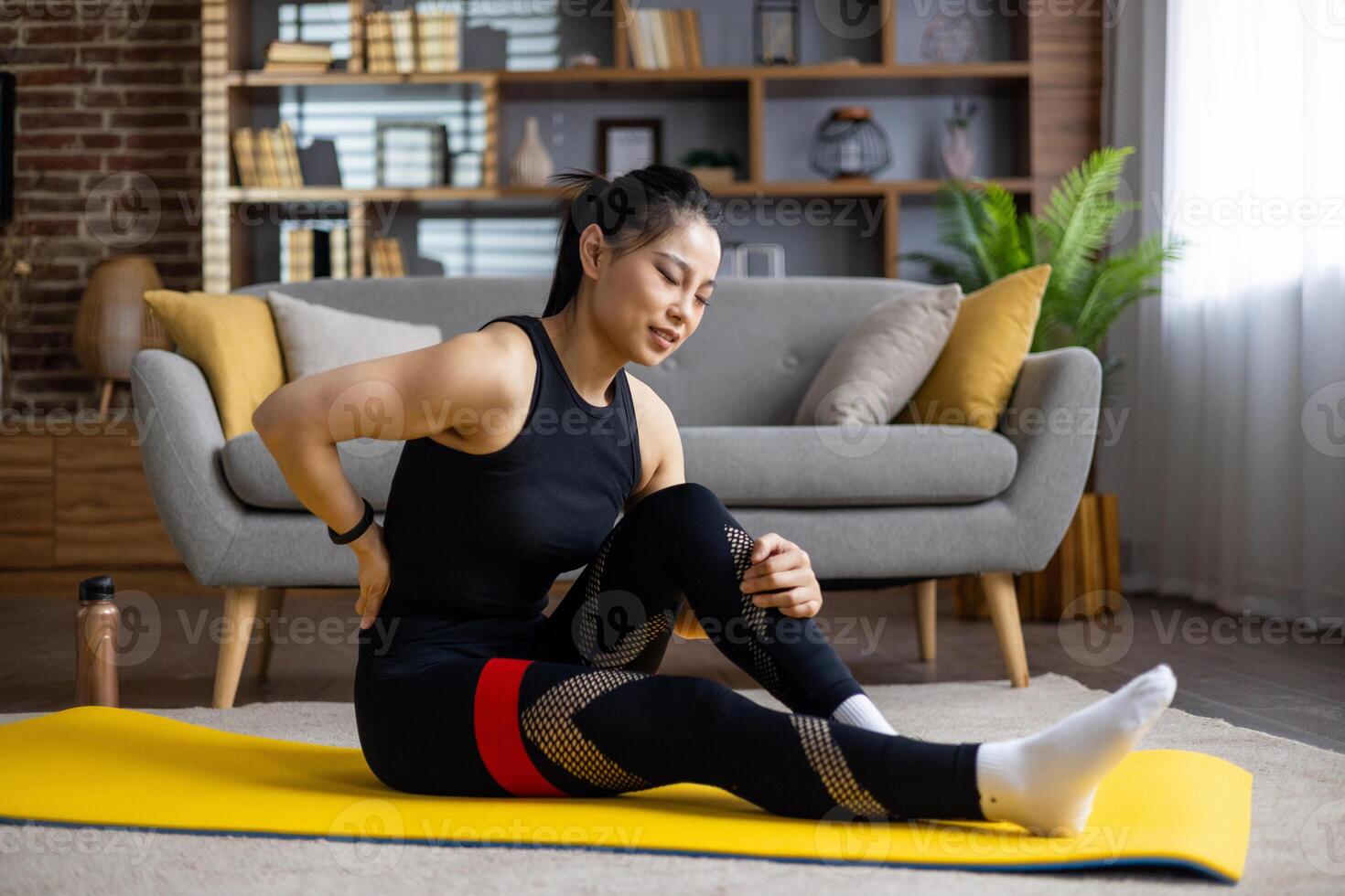 Asian woman experiencing back pain sitting on yoga mat in living room. Wearing black workout clothes, gently stretching for relief. Focus on health and relaxation indoors. photo