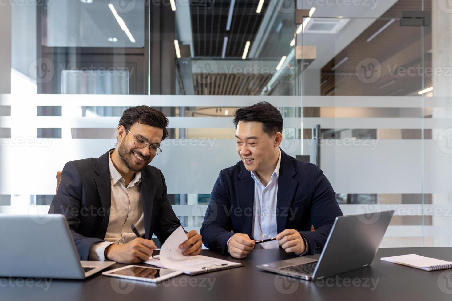 Two professionals are working together, reviewing documents in a modern conference room setting. They are surrounded by laptops and other tools, engaging in a discussion. photo